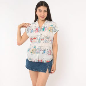 May include: A short-sleeved button-down shirt in white featuring a colorful, hand-drawn style print of various buildings and architectural structures in shades of red, blue, yellow, and green, paired with a dark blue denim mini skirt with a side slit.