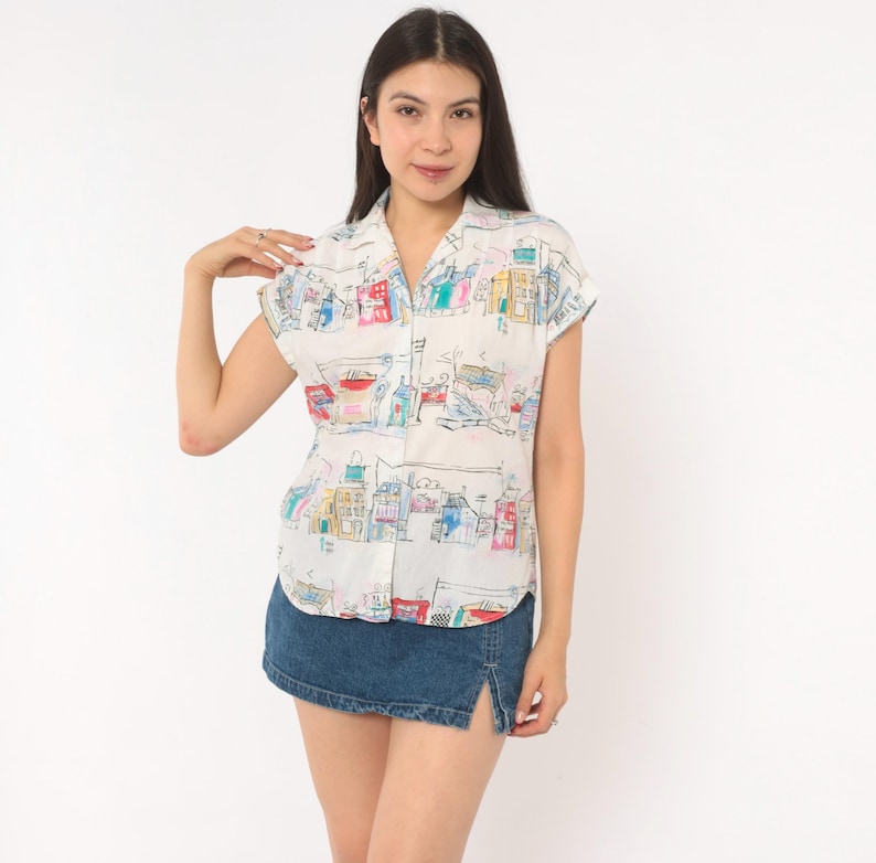 May include: A short-sleeved button-down shirt in white featuring a colorful, hand-drawn style print of various buildings and architectural structures in shades of red, blue, yellow, and green, paired with a dark blue denim mini skirt with a side slit.