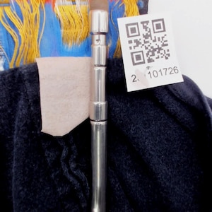 May include: Close-up of a dark blue garment with a silver metal rod in the center. A white tag with a QR code and the number "2. 101726" is attached. The garment has colorful accents of blue, yellow, and red.