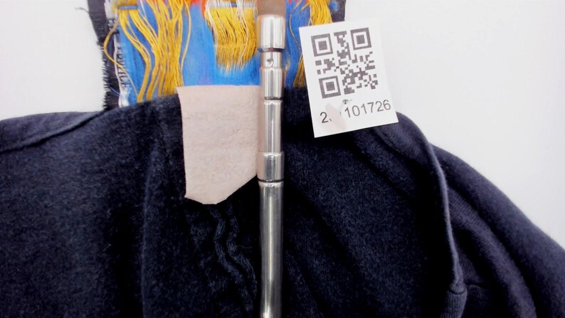 May include: Close-up of a dark blue garment with a silver metal rod in the center. A white tag with a QR code and the number "2. 101726" is attached. The garment has colorful accents of blue, yellow, and red.