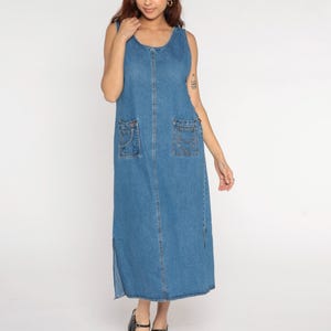 Könnte beinhalten: Ein ärmelloses, hellblaues Jeanskleid mit zwei Vordertaschen und Seitenschlitzen. Das Kleid hat einen runden Ausschnitt und ein bodenlanges Design. Das Model trägt schwarze Sandalen.
