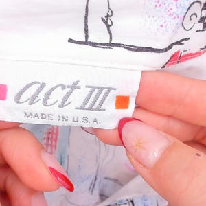 May include: A white clothing label with the text act III in a grey font, flanked by a small pink square on the left and an orange square on the right. Below the brand name, the text reads MADE IN U.S.A. The label is attached to a white garment.