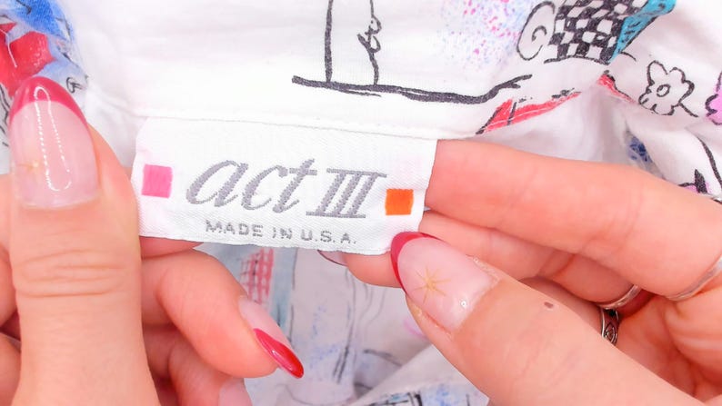 May include: A white clothing label with the text act III in a grey font, flanked by a small pink square on the left and an orange square on the right. Below the brand name, the text reads MADE IN U.S.A. The label is attached to a white garment.