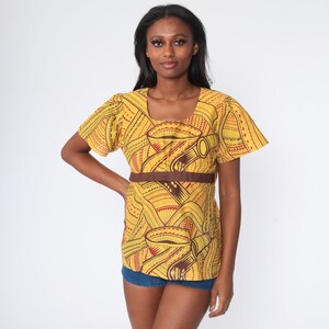 May include: Yellow top with a brown waist band and short sleeves. The top has a repeating pattern in red and brown. The neckline is square. The model is wearing blue jean shorts.