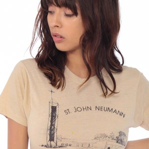 May include: Beige short-sleeved t-shirt featuring a black and white illustration of a building with a cross on top and the text "ST. JOHN NEUMANN". The shirt is made of a soft, light-colored fabric.