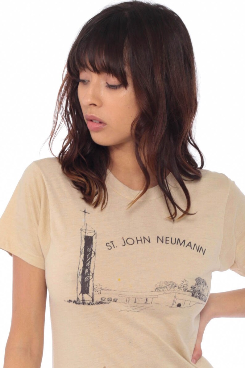 May include: Beige short-sleeved t-shirt featuring a black and white illustration of a building with a cross on top and the text "ST. JOHN NEUMANN". The shirt is made of a soft, light-colored fabric.