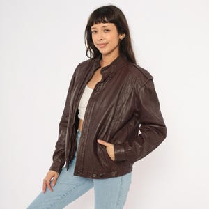 May include: A dark brown leather jacket with a stand-up collar and a zipper closure. The jacket has a classic bomber style with pockets and a slightly oversized fit. The model is wearing light blue jeans.