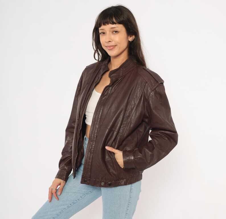 May include: A dark brown leather jacket with a stand-up collar and a zipper closure. The jacket has a classic bomber style with pockets and a slightly oversized fit. The model is wearing light blue jeans.