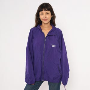 May include: A purple Reebok windbreaker jacket with a full zipper closure. The jacket has a small white Reebok logo on the chest. The jacket is made of a lightweight material and has a collar.