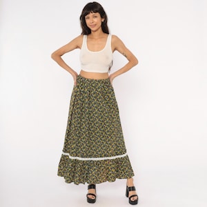 Puede incluir: Falda larga con estampado floral y ribete de encaje blanco en el dobladillo. La falda presenta un fondo oscuro con pequeñas flores amarillas y blancas. La modelo lleva una camiseta sin mangas blanca y sandalias de plataforma negras.