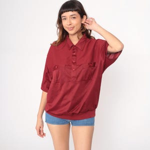Puede incluir: Una camisa burdeos de manga corta con cuello y tapeta de botones. La camisa tiene dos bolsillos en el pecho y un dobladillo fruncido. La modelo lleva pantalones cortos vaqueros azules.