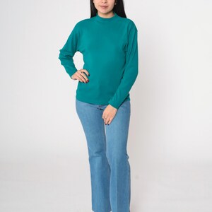 Pode incluir: Uma camisa de manga comprida verde-azulado e cal&ccedil;as de ganga flare azul claro. A camisa tem gola alta. As cal&ccedil;as de ganga s&atilde;o de ganga clara. A pessoa est&aacute; a usar sand&aacute;lias brancas com plataforma de madeira.