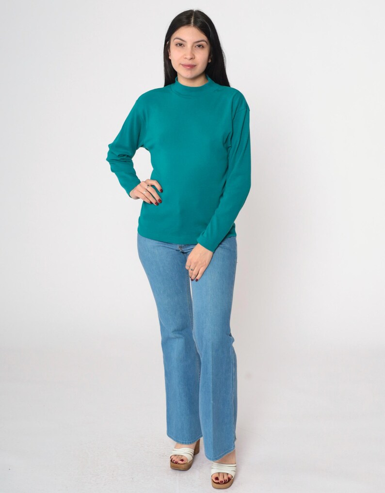 Pode incluir: Uma camisa de manga comprida verde-azulado e cal&ccedil;as de ganga flare azul claro. A camisa tem gola alta. As cal&ccedil;as de ganga s&atilde;o de ganga clara. A pessoa est&aacute; a usar sand&aacute;lias brancas com plataforma de madeira.
