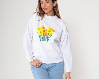 May include: White sweatshirt featuring a floral design of yellow and orange flowers with green stems. The crew neck sweatshirt is paired with blue jeans. The floral design is centered on the chest.