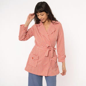 May include: A red and white gingham patterned jacket with a tie waist and two front pockets. The jacket has a collar and three-quarter length sleeves. The model is wearing blue jeans.