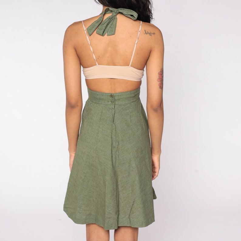 May include: Olive green halter dress with a high waist and a-line skirt. The dress features a halter tie and a backless design. The dress is made of a textured fabric and has a button closure at the back.