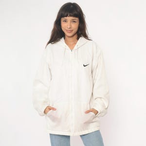 Puede incluir: Chaqueta blanca con capucha, cierre de cremallera y un peque&ntilde;o logotipo negro de Nike en el pecho izquierdo. La chaqueta tiene dos bolsillos delanteros y se lleva sobre unos vaqueros azules. La modelo sonr&iacute;e.