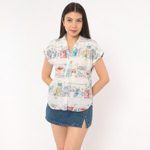 May include: A short-sleeved white button-down shirt featuring a colorful hand-drawn style print of various buildings and city structures in shades of red, blue, yellow, and green, paired with a dark blue denim mini skirt with a front slit.