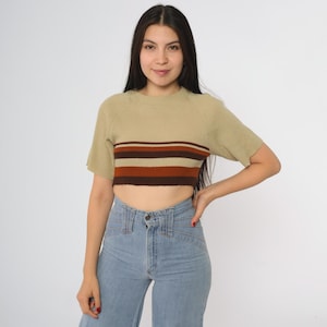 May include: A short-sleeved, cropped top in a light tan color with a horizontal stripe design in brown and orange. The top is paired with light blue denim jeans.