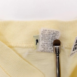 May include: Close-up of a pale yellow V-neck shirt. The shirt has a ribbed collar and a tag with care instructions and a QR code. A stylus is in the foreground.