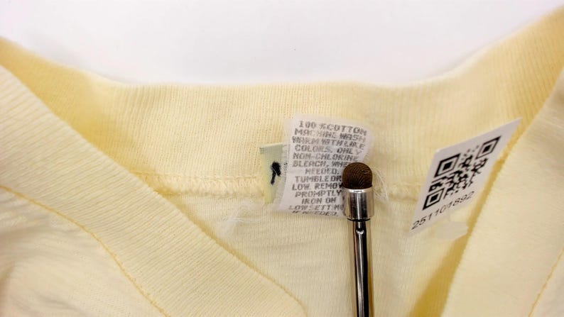 May include: Close-up of a pale yellow V-neck shirt. The shirt has a ribbed collar and a tag with care instructions and a QR code. A stylus is in the foreground.