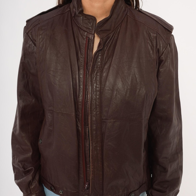 May include: A brown leather jacket with a zip-up front and a stand-up collar. The jacket has shoulder epaulets and a classic, vintage style. The leather appears to be slightly distressed, adding to its character.