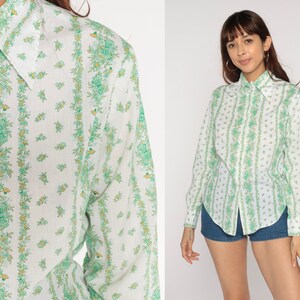 Puede incluir: Una camisa blanca de manga larga con botones y estampado floral verde y blanco. La camisa tiene un cuello puntiagudo y cierre de botones en la parte delantera.