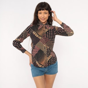 May include: A long-sleeve button-down shirt featuring a black base with a multi-patterned design in shades of gold, pink, and white. The shirt displays a mix of geometric shapes, polka dots, and paisley motifs across the fabric and collar.