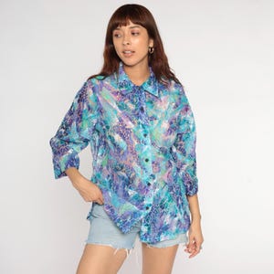 Op de afbeelding: Een overhemd met knopen met een levendig, abstract patroon in tinten blauw, paars en teal. De blouse heeft een kraag en driekwart mouwen. De stof lijkt licht en semi-transparant. De blouse wordt gecombineerd met een spijkerbroekshort.