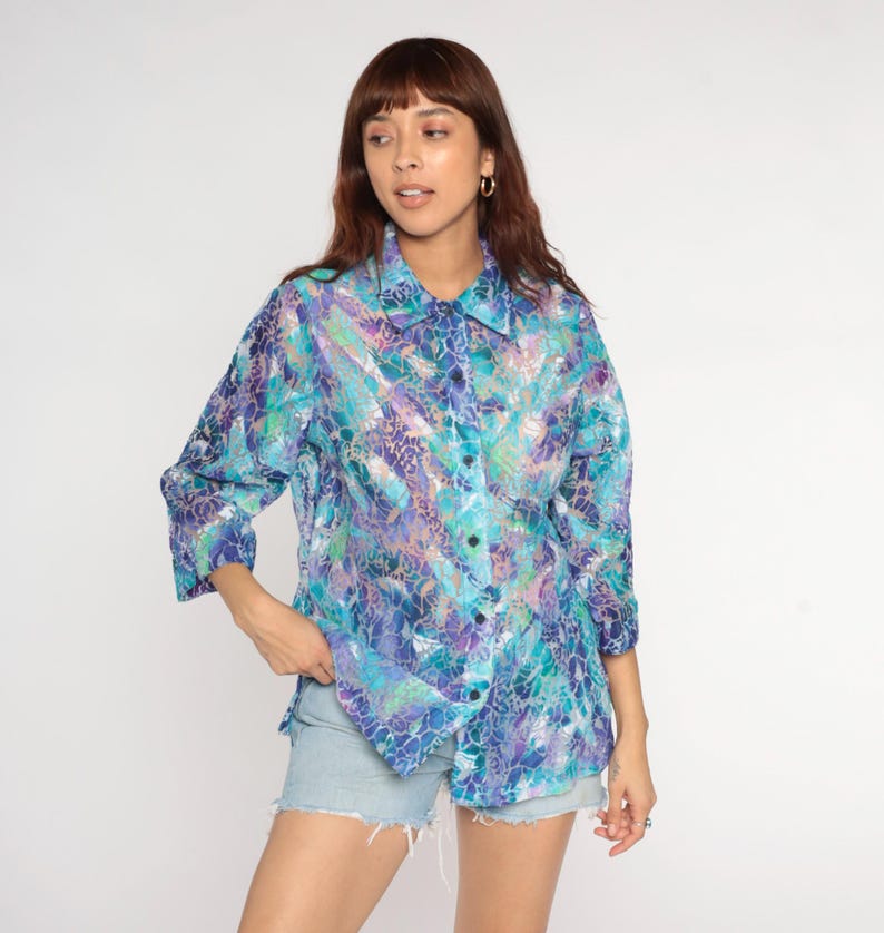 Op de afbeelding: Een overhemd met knopen met een levendig, abstract patroon in tinten blauw, paars en teal. De blouse heeft een kraag en driekwart mouwen. De stof lijkt licht en semi-transparant. De blouse wordt gecombineerd met een spijkerbroekshort.