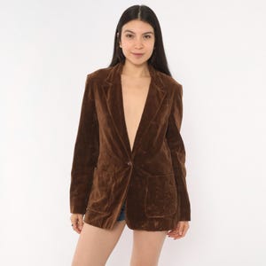 May include: A dark brown velvet blazer featuring a single-button closure, notched lapels, and two large patch pockets on the front. The jacket has long sleeves and a structured fit, crafted from a soft, light-reflecting velvet fabric.