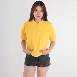 May include: A yellow short-sleeved polo shirt with a button-up collar and a pocket on the left side. The shirt is tucked into a pair of black denim shorts.