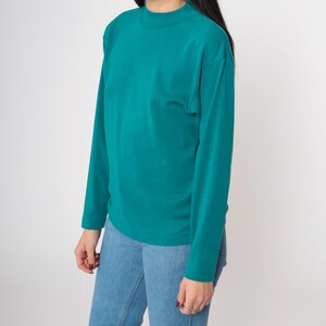 Pode incluir: Camiseta de manga comprida verde-azulado com gola alta. A camisa &eacute; combinada com jeans azul claro. A modelo est&aacute; usando a camisa e jeans em um fundo branco. A camisa tem um design simples e cl&aacute;ssico.