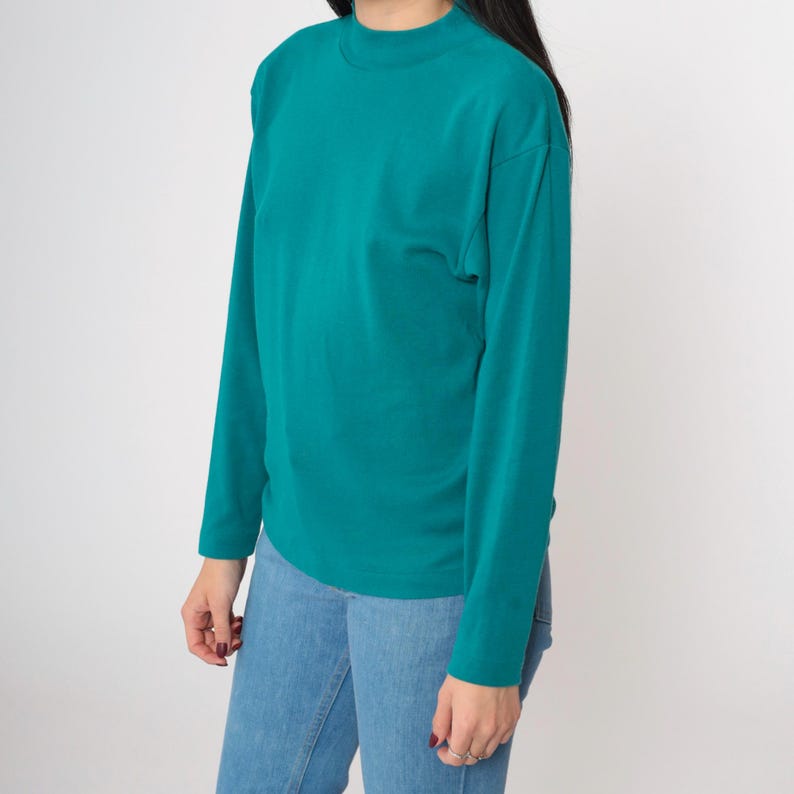 Pode incluir: Camiseta de manga comprida verde-azulado com gola alta. A camisa &eacute; combinada com jeans azul claro. A modelo est&aacute; usando a camisa e jeans em um fundo branco. A camisa tem um design simples e cl&aacute;ssico.