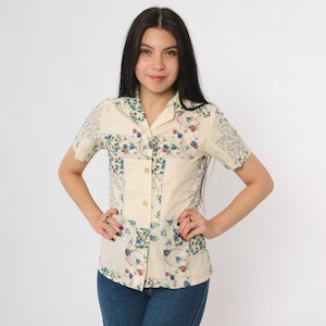 Può includere: Camicia color crema a maniche corte con un design patchwork. La camicia presenta motivi floreali blu, rossi e verdi, con colletto e bottoni in legno. La camicia è abbinata a jeans blu.