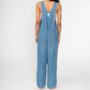 May include: A pair of light blue denim dungarees with a wide leg and a V-neck. The dungarees have a small green triangle logo on the front.