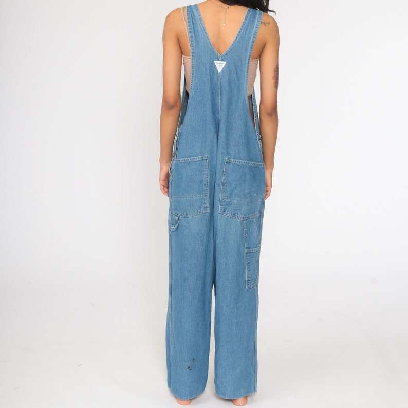 May include: A pair of light blue denim dungarees with a wide leg and a V-neck. The dungarees have a small green triangle logo on the front.