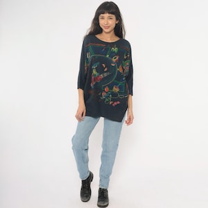 May include: A dark blue, oversized top with a colorful, abstract design. The top has three-quarter sleeves and a round neckline. The person is wearing light blue jeans and black boots.