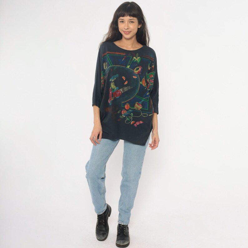 May include: A dark blue, oversized top with a colorful, abstract design. The top has three-quarter sleeves and a round neckline. The person is wearing light blue jeans and black boots.