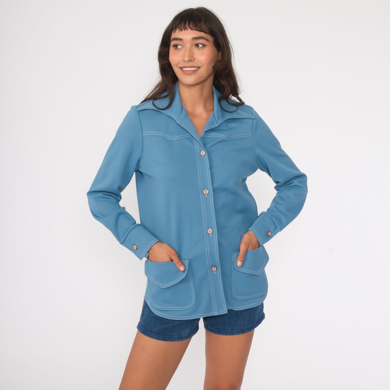 May include: A light blue long-sleeved shirt with a collared neckline, button closure, and two patch pockets. The shirt has white stitching details.