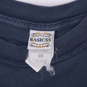 May include: A dark blue t-shirt with a white tag that reads "Gold Basicss Label" and "Fabrique aux Mexique" with instructions to see the reverse side for care instructions. The tag also includes a size label that reads "XL" and "100% Ring Spun Cotton Made in Mexico Care on Reverse".