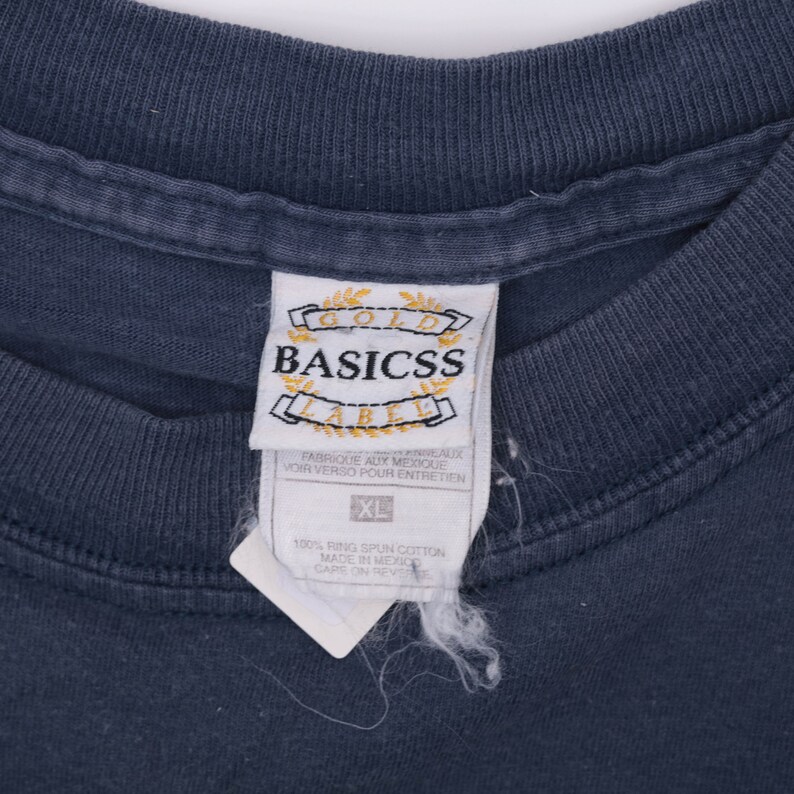 May include: A dark blue t-shirt with a white tag that reads "Gold Basicss Label" and "Fabrique aux Mexique" with instructions to see the reverse side for care instructions. The tag also includes a size label that reads "XL" and "100% Ring Spun Cotton Made in Mexico Care on Reverse".