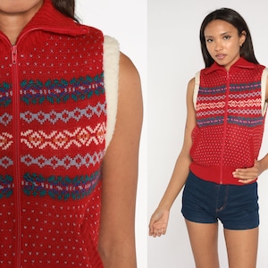 May include: Red knitted waistcoat with a white faux fur collar and a zip fastening. The waistcoat has a geometric pattern in shades of blue, green, and pink.