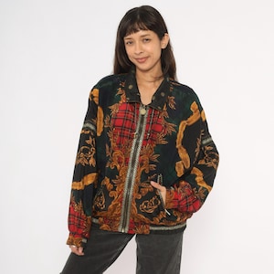 May include: A vintage bomber jacket with a vibrant mix of patterns. The jacket features a red plaid design, gold baroque-style accents, and a black background. It has a full zipper, a collar, and a pocket. The jacket is a statement piece.