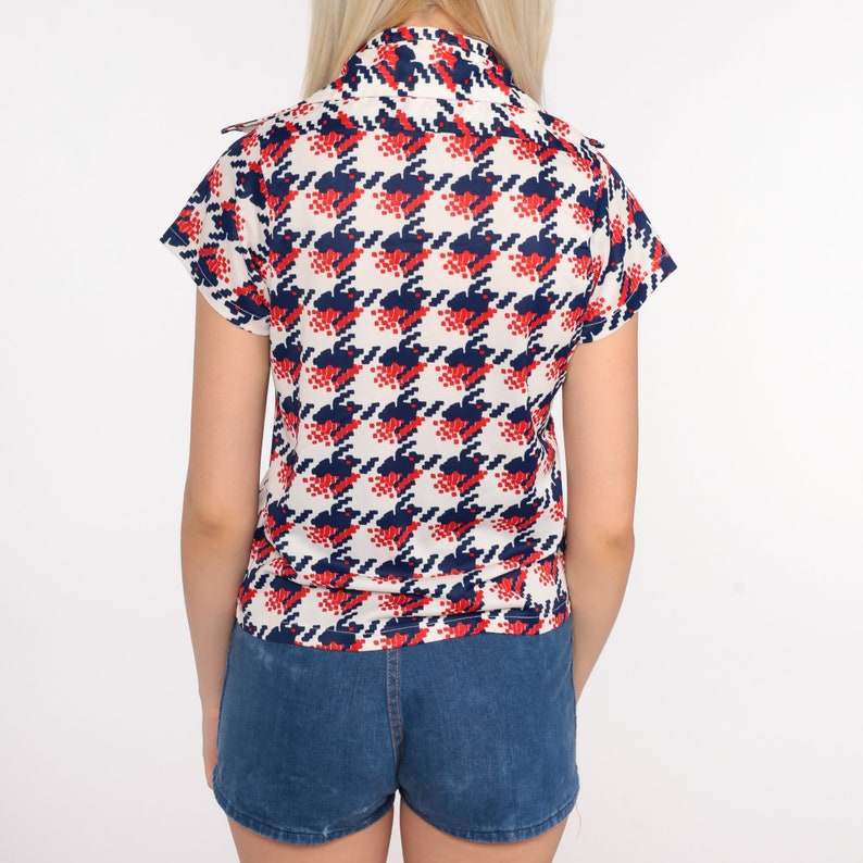 May include: A white short-sleeved shirt with a red and blue houndstooth pattern. The shirt has a collar and a button-up front. The model is wearing blue denim shorts.