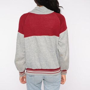 Op de afbeelding: Een grijs en bordeauxkleurig colorblock sweatshirt. Het bovenste gedeelte is bordeauxrood, met een witte streep die het scheidt van de grijze body. De manchetten en zoom hebben bordeauxrode en witte strepen. De kraag is een col.