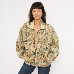 Puede incluir: Chaqueta vintage con cremallera y estampado abstracto en tonos verdes, beige y marrón. La chaqueta tiene cuello, bolsillos y cremallera marrón oscuro. La modelo lleva vaqueros azules.