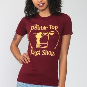 May include: A maroon t-shirt with a gold graphic of a hand throwing a dart at a dartboard. The graphic says "the Double Top Dart Shop".