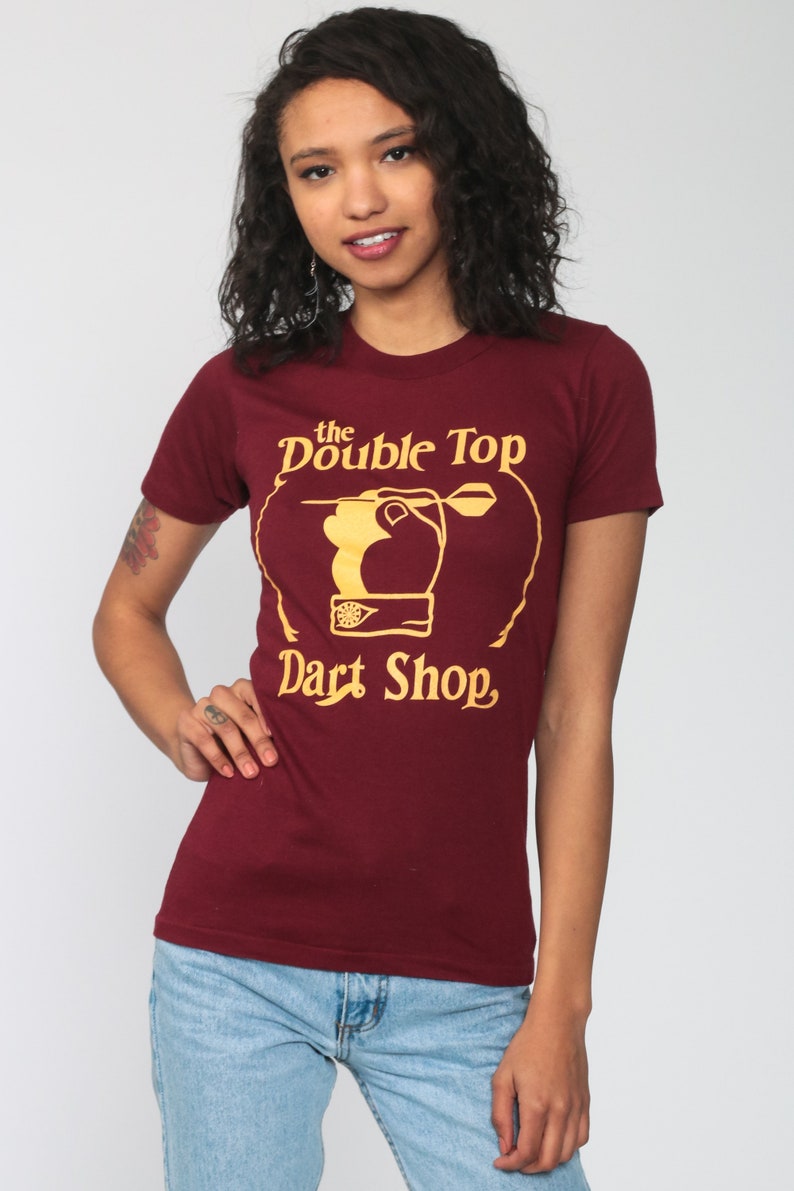 May include: A maroon t-shirt with a gold graphic of a hand throwing a dart at a dartboard. The graphic says "the Double Top Dart Shop".