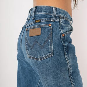 Puede incluir: Vaqueros de mezclilla azules con un dise&ntilde;o cl&aacute;sico de cinco bolsillos. Los vaqueros presentan un parche de cuero marr&oacute;n con el logotipo "Wrangler" en el bolsillo trasero. Los vaqueros tienen remaches de cobre y cierre de bot&oacute;n.